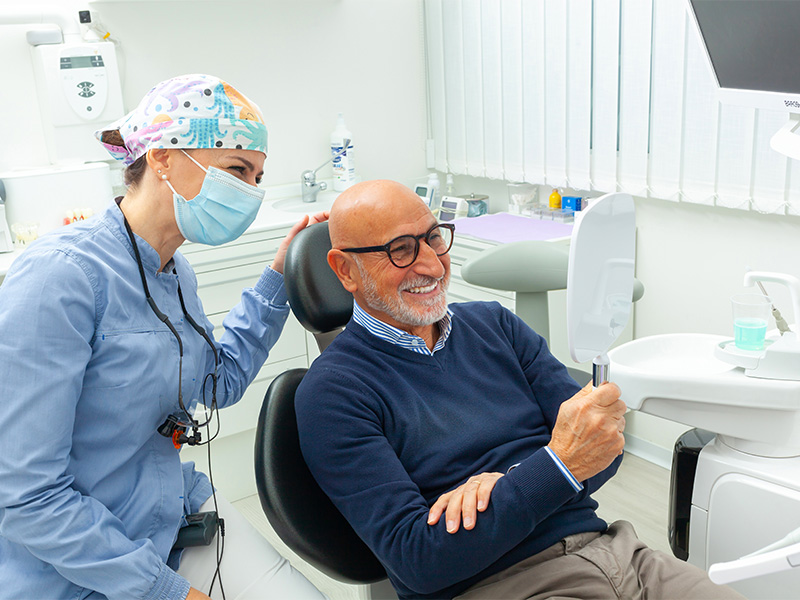 Zahnärztin zeigt älterem Herrn das Behandlungsergebnis. Beide lächeln. Moderne Praxis für Oralchirurgie in Lübeck.